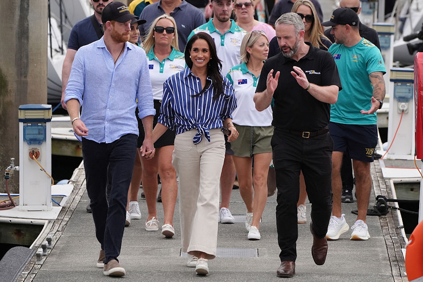 Prince Harry and Meghan Markle holding hands while walking along a marina dock, with a man in a black polo gesturing beside them and a group of people following behind.