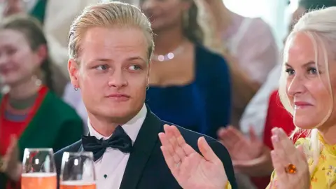 Marius Borg Høiby in a tuxedo with bow tie seated next to Crown Princess Mette-Marit, who is applauding, at a formal event with glasses of beverage on the table.