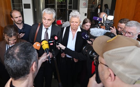 Two prosecutors, a man in a suit and tie and a woman in a black blazer, speaking to a crowd of reporters holding microphones and cameras outside a building.
