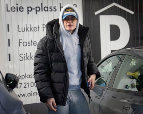 Marius Borg Høiby wearing a black puffer jacket, white hoodie, and cap stands next to a gray car in front of a white building with text.