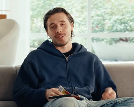 Brooklyn Beckham in a dark blue hoodie sits on a gray couch holding a remote control, with windows behind him.