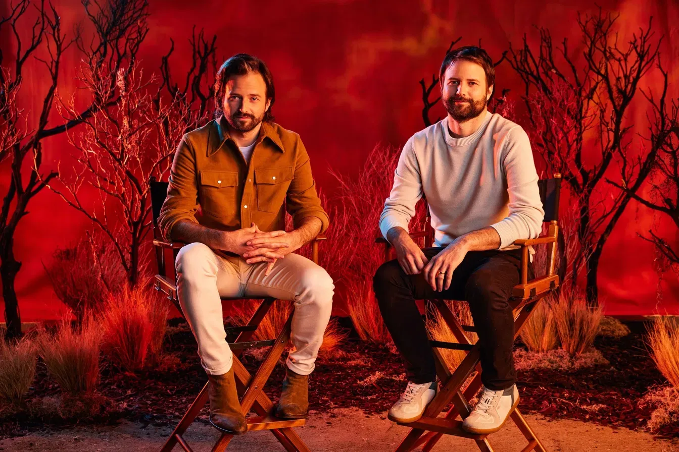 Matt and Ross Duffer seated in director's chairs against a red-lit backdrop with bare black trees, one in a brown shirt jacket and white pants, the other in a cream sweater and dark pants.