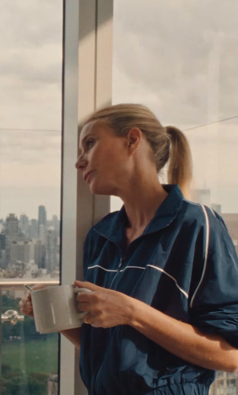 Gwyneth Paltrow in a dark blue shirt holds a white mug while gazing out a high-rise window at a cityscape below.