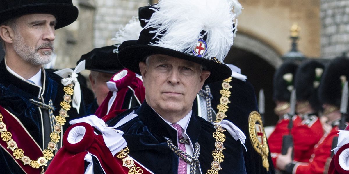 Prince Andrew in formal ceremonial regalia featuring a black hat with white plumes, ornate gold trim, and red velvet robes stands solemnly during an official state occasion.