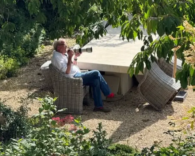 Jeremy Clarkson in a white shirt and jeans sits in a wicker chair photographing in a shaded garden area surrounded by plants.