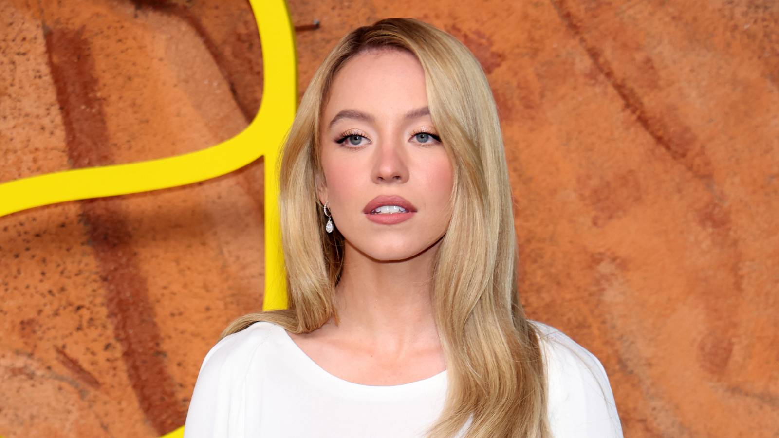 Sydney Sweeney with blonde hair wearing a white top stands against an orange textured backdrop with yellow curved design elements.
