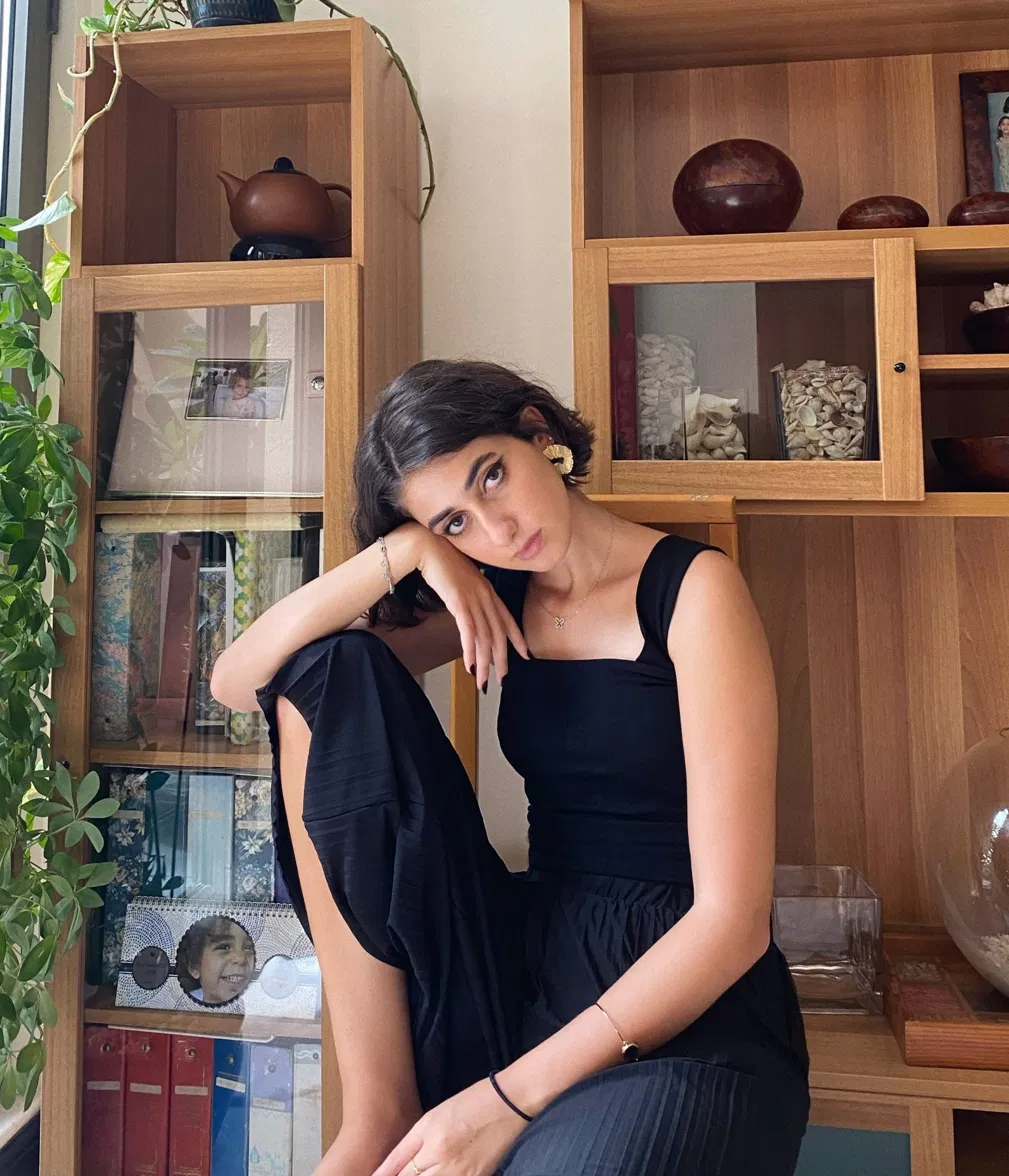Rama Duwaji in a black sleeveless top with gold fan earrings and a delicate necklace, seated in front of wooden shelving units filled with books, ceramics, and plants.