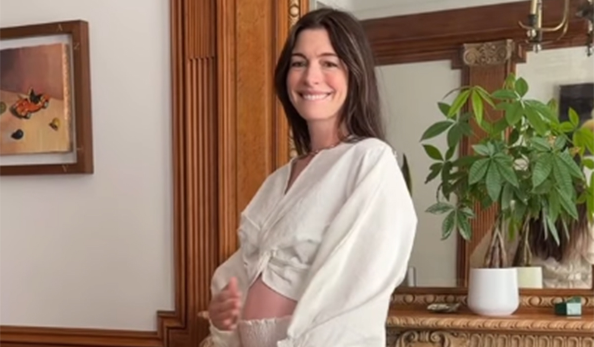 Anne Hathaway smiling and showing her pregnant belly in a white wrap top, standing indoors in front of a wooden mantel with a potted plant and framed artwork.