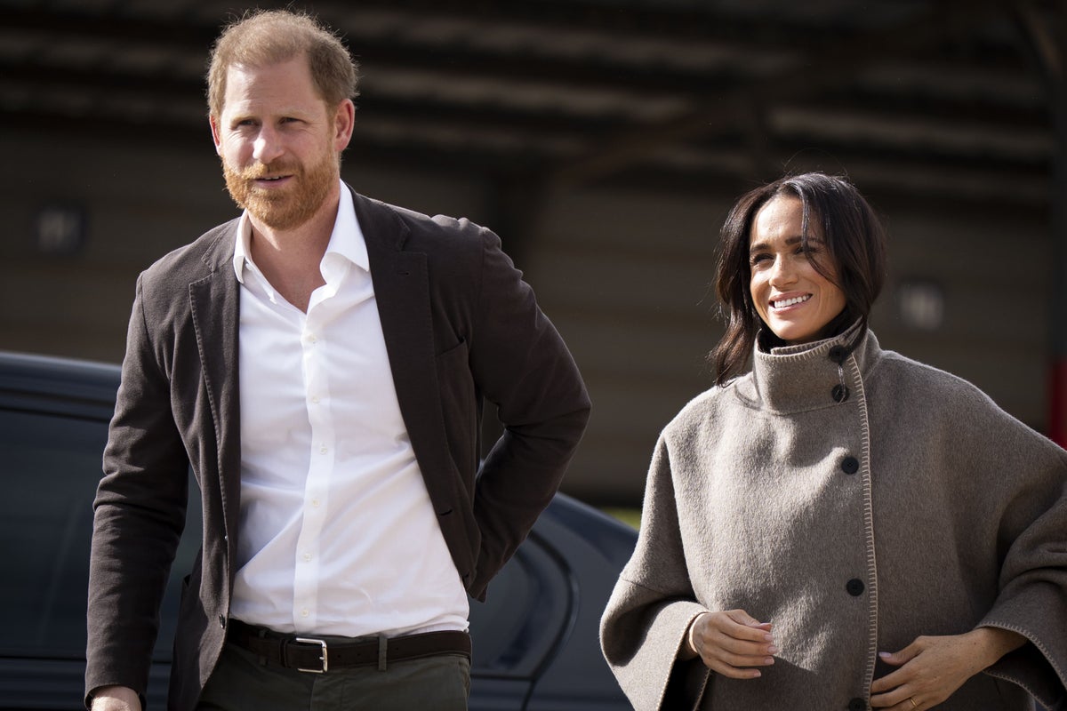 Prince Harry and Meghan Markle walking together outdoors, Harry in dark blazer and white shirt, Meghan in beige turtleneck sweater, both smiling.