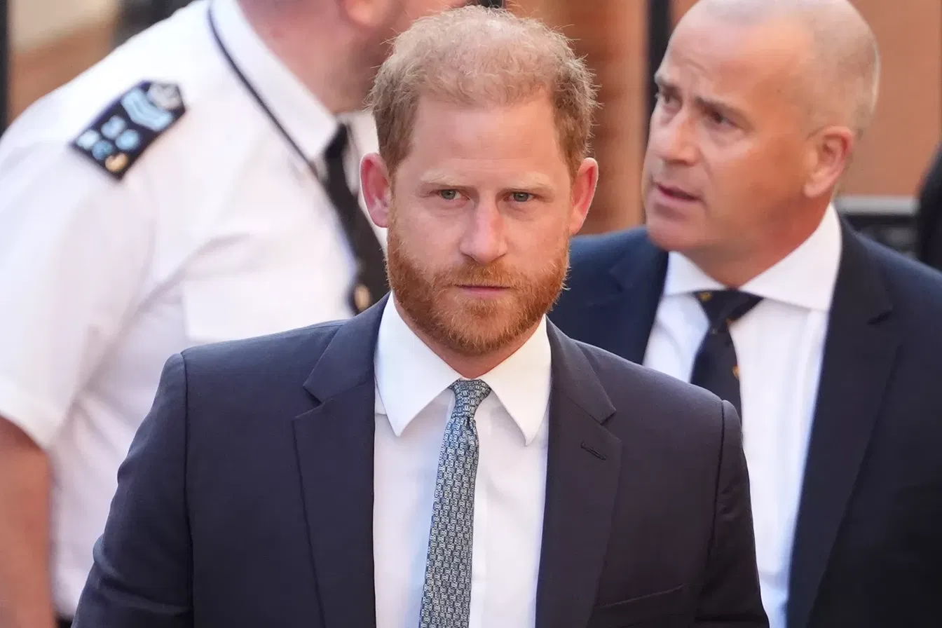 Prince Harry in a navy suit and patterned tie walking outdoors, flanked by a uniformed officer and a man in a dark suit.