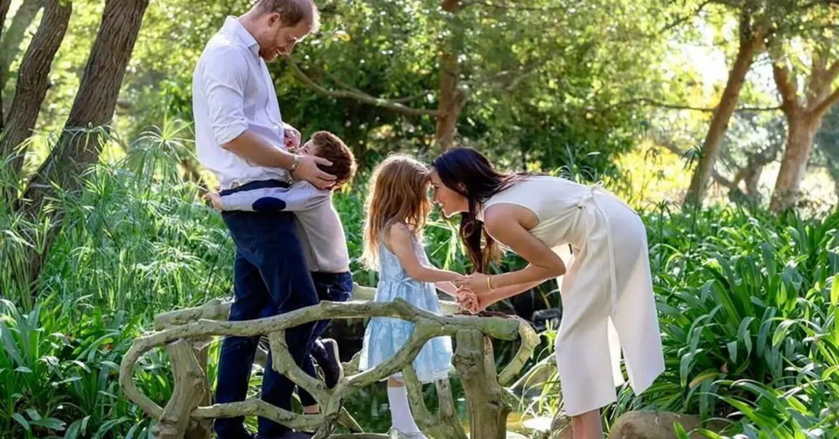 Prince Harry holding Archie and Meghan Markle bending down toward Lilibet on a wooden bridge surrounded by greenery, with Meghan wearing a cream sleeveless dress.