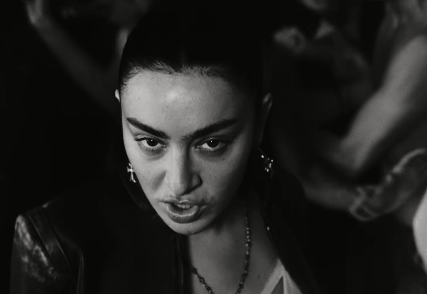 Charli XCX in dark clothing with pearl earrings gazes downward in this black and white portrait photograph.