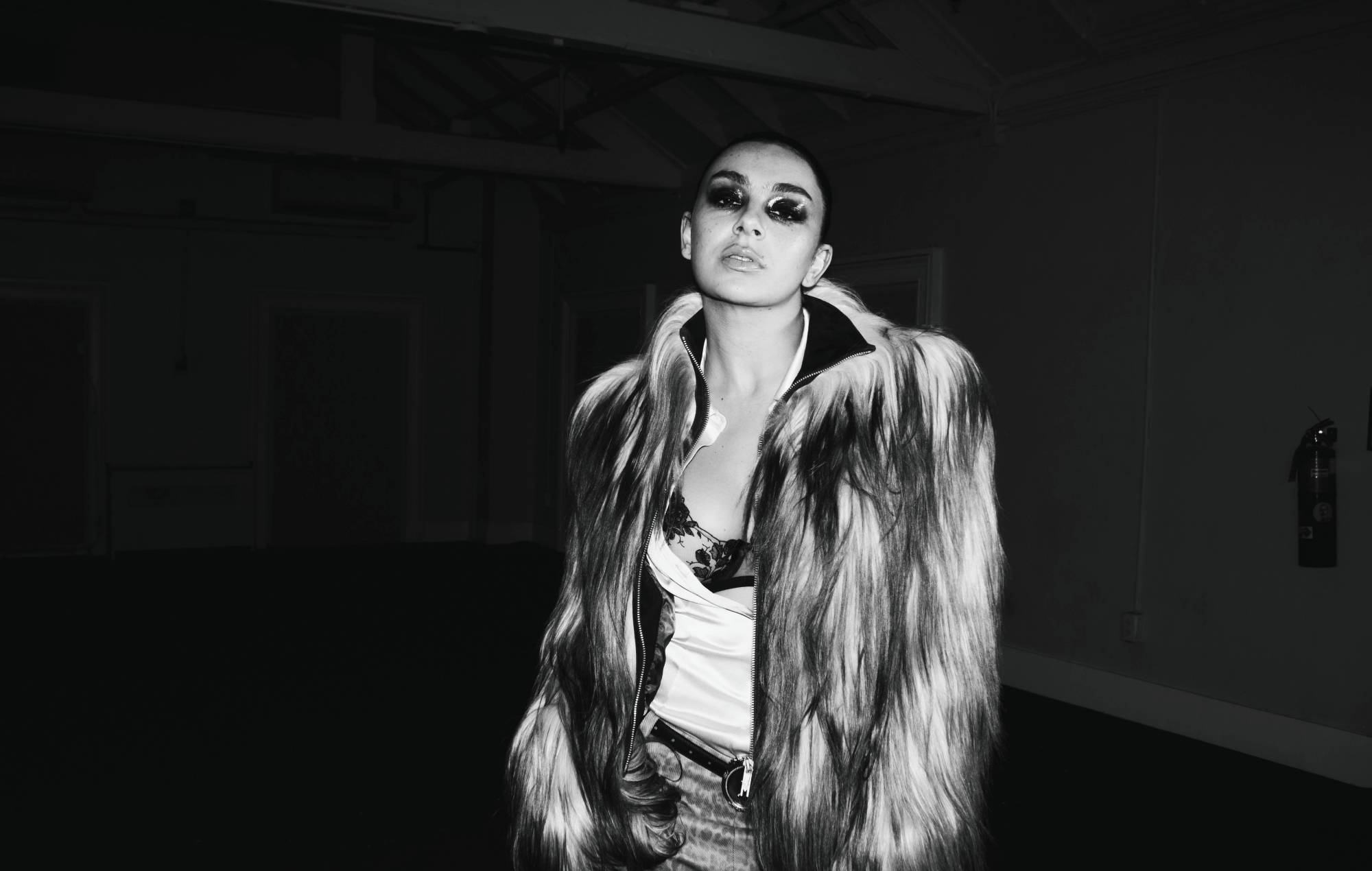 Charli XCX wearing a textured fur coat and bold eye makeup stands in a dimly lit interior space, looking upward.