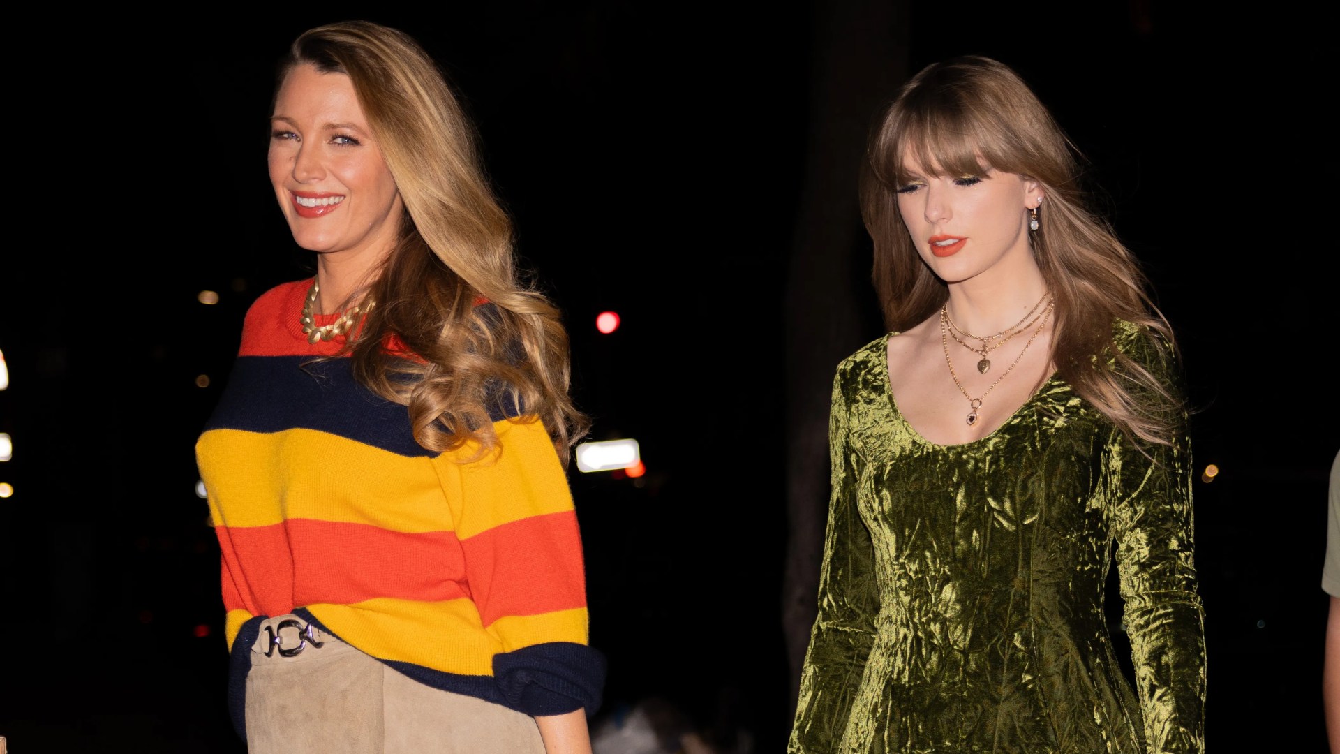 Blake Lively and Taylor Swift standing together at night; Blake on left wears colorful striped sweater, Swift on right wears green velvet top.