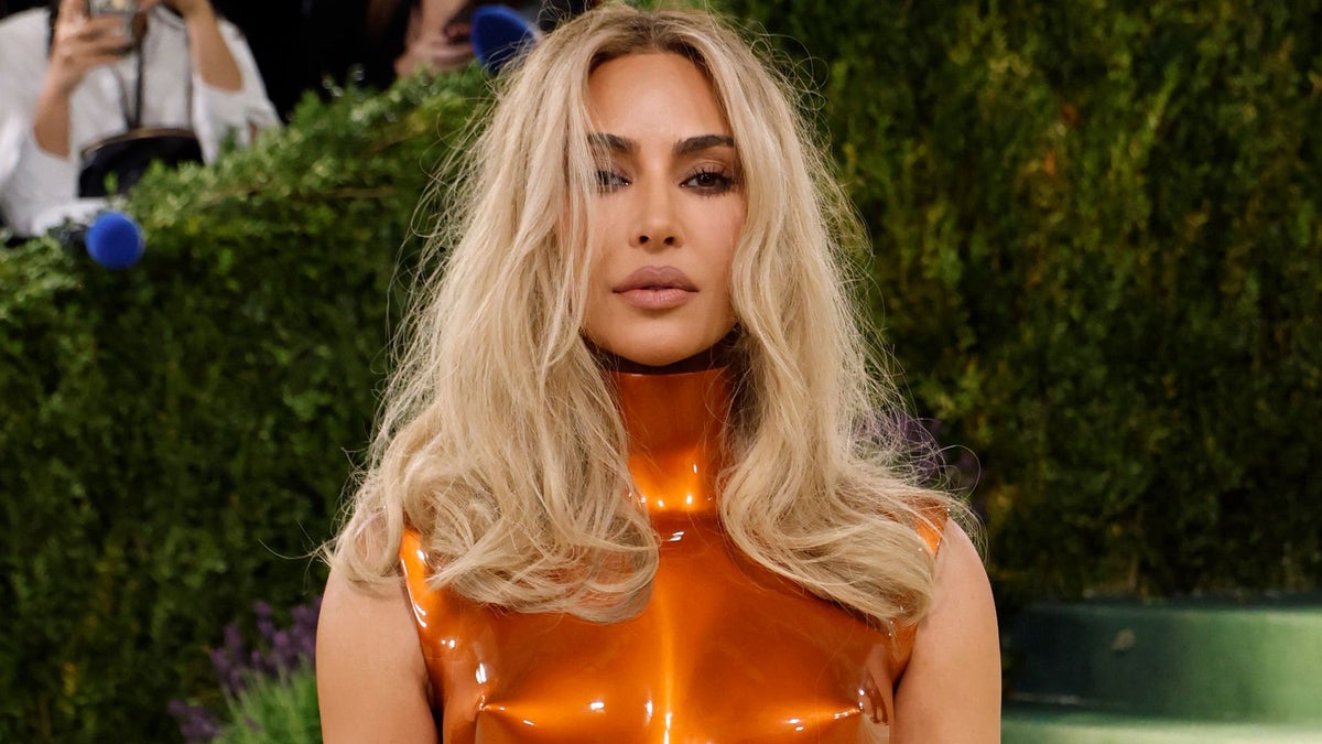 Kim Kardashian with long platinum blonde hair wearing a glossy orange sleeveless top stands against a green foliage backdrop.