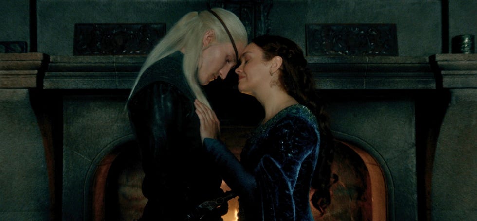 Two characters in a dimly lit stone room with a fireplace, one in black with long white hair leaning close to the other in a dark blue gown with dark hair.