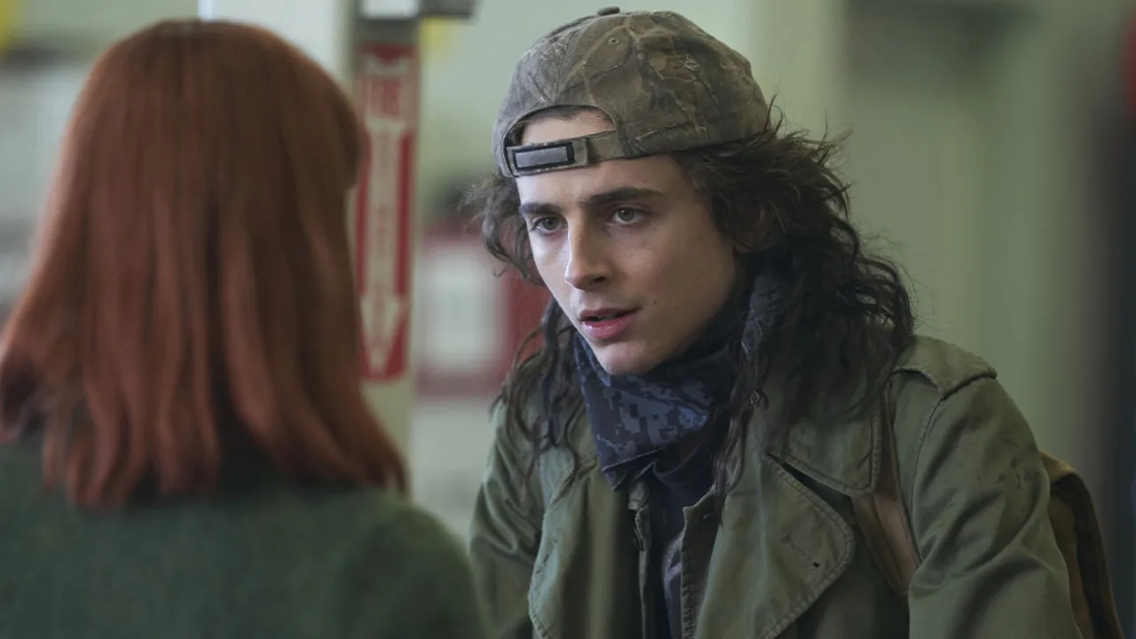 A young man with long dark hair in a backwards camo cap and olive jacket with a blue bandana around his neck, talking to a red-haired woman in a dimly lit indoor setting.