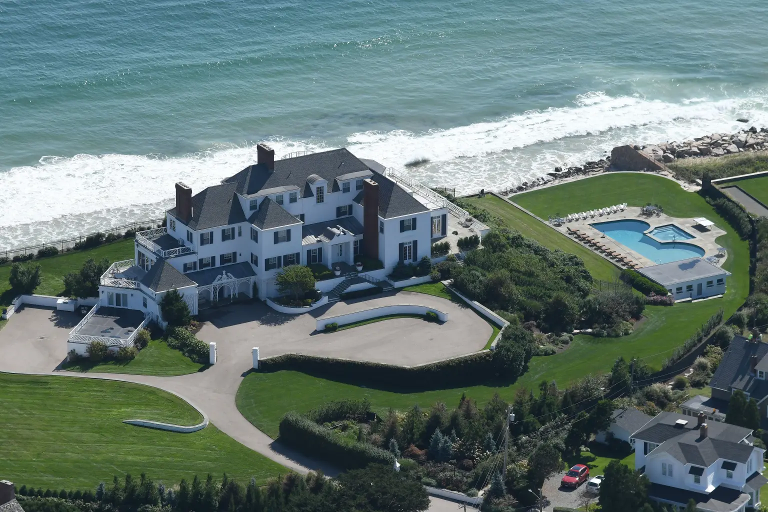 Aerial view of a large white Colonial-style mansion on a seafront estate with a pool, manicured lawns, and a private driveway, with the ocean directly behind.
