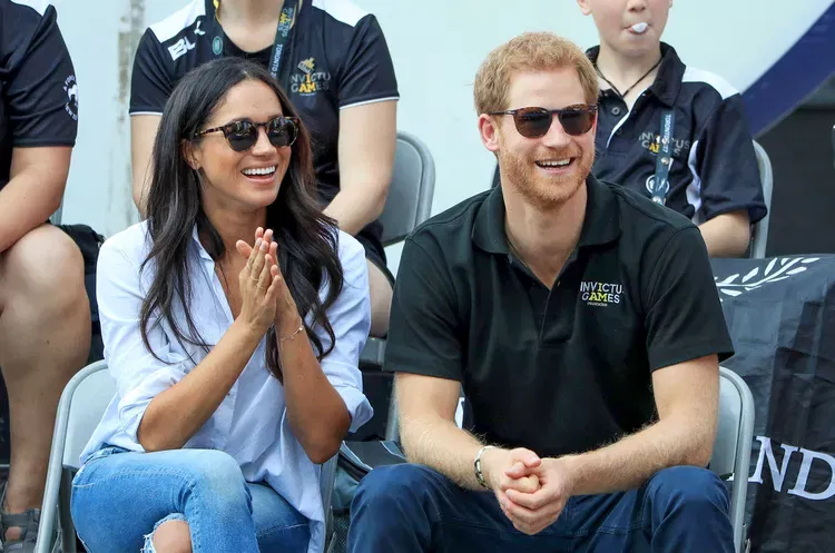 A woman in a white shirt and jeans and a man in an Invictus Games polo shirt, both wearing sunglasses and seated together at a sporting event.