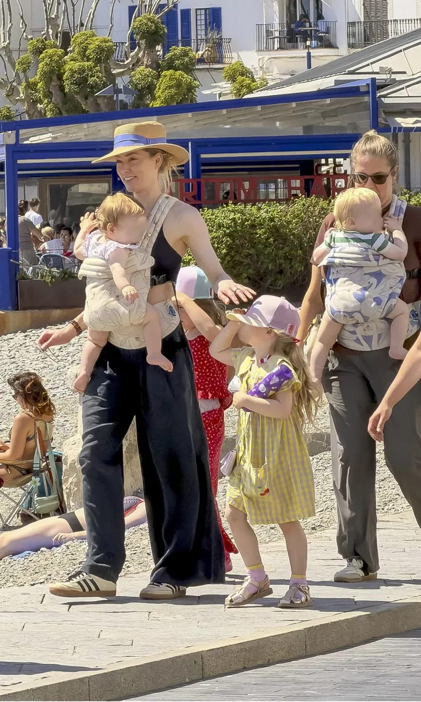 A woman in a straw hat, black top and wide-leg trousers carrying a baby in a front carrier, with a young girl in a yellow dress beside her and another woman carrying a baby behind them, on a sunny seafront promenade.