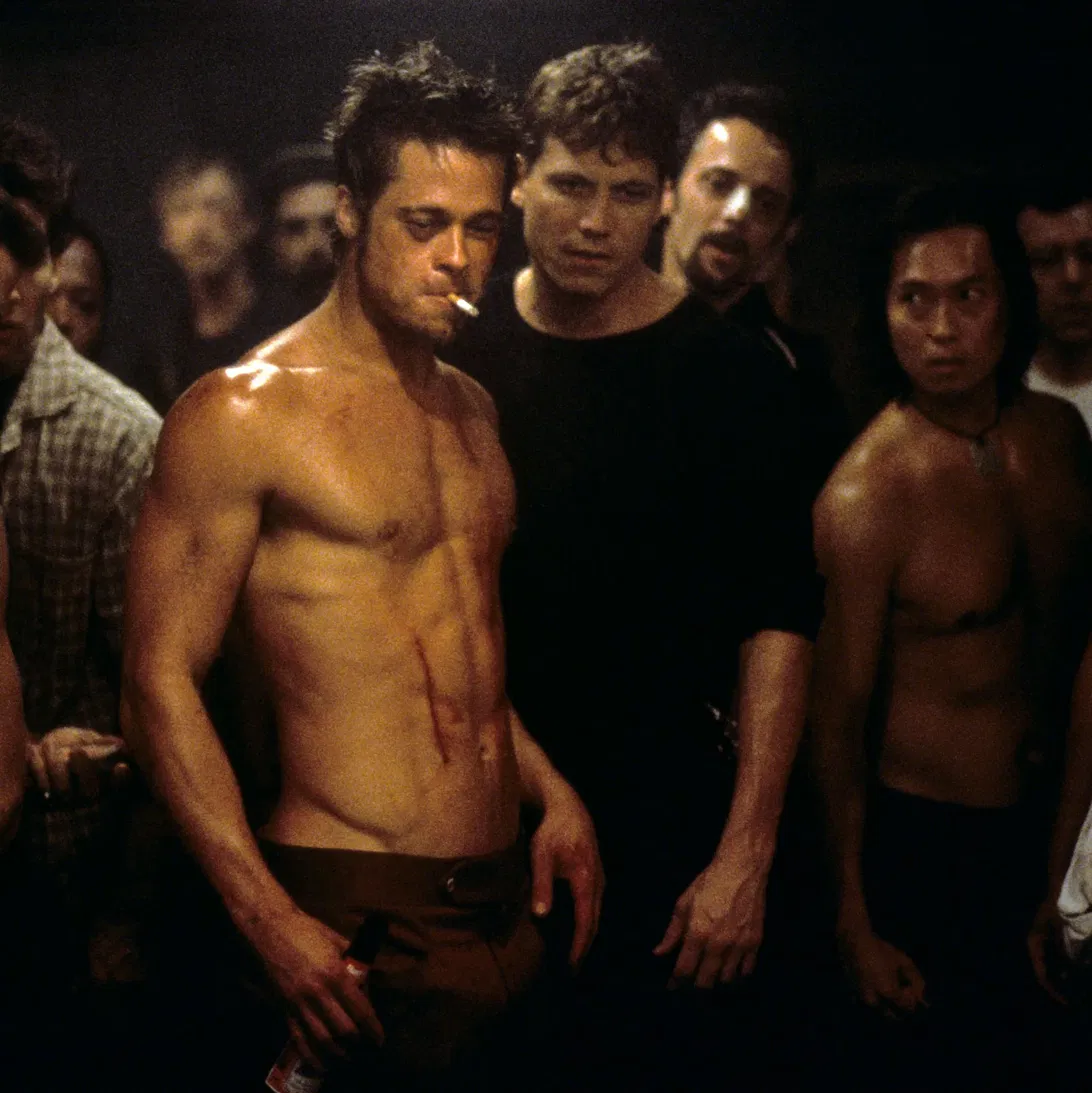 A shirtless man with a cigarette in his mouth, visible cuts on his torso, stands in front of a crowd of men in a dimly lit basement setting.