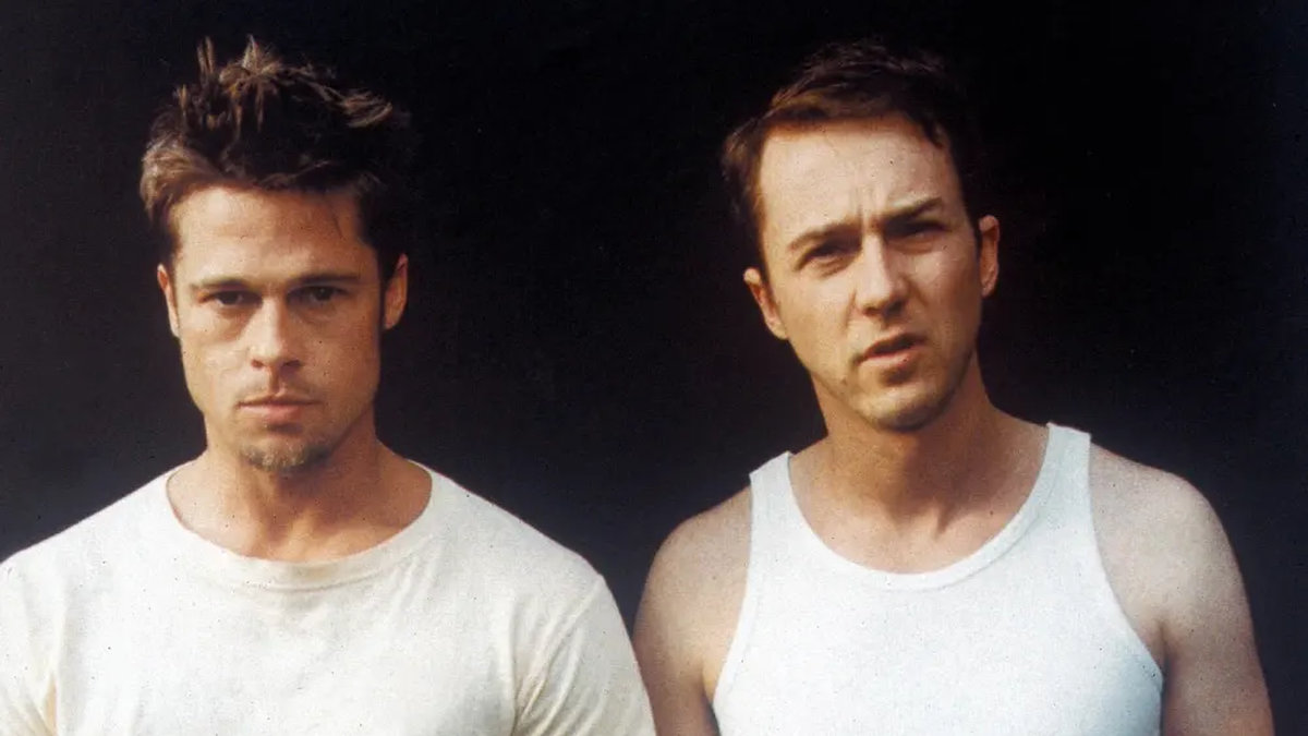 Two men in white tops stand side by side against a dark background — one in a crew-neck t-shirt and the other in a sleeveless vest 