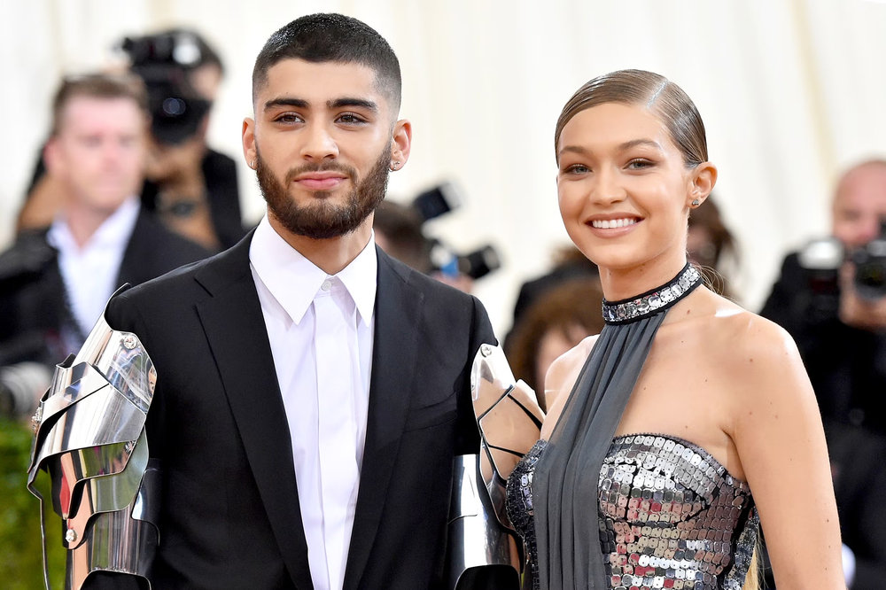 A man in a black suit with silver armour panels at the wrists stands beside a woman in a silver sequined halter gown, both on a red carpet.