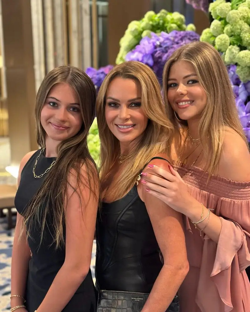 Three women with long brown hair pose together in front of a floral arrangement — the woman in the centre wearing a black leather dress, flanked by two younger women in black and pink respectively.