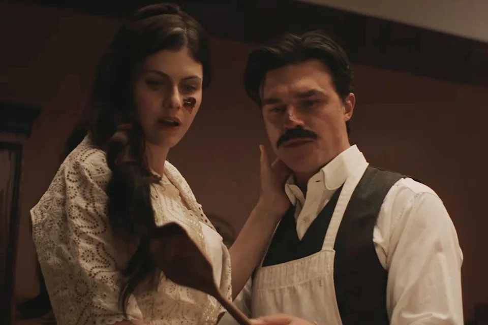 A woman in a lace-trimmed blouse and a man with a mustache wearing a vest and apron stand together indoors.