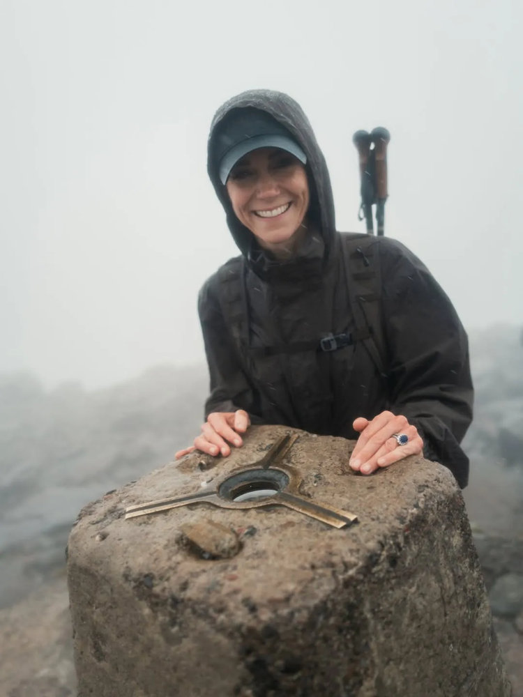 A woman in a black waterproof jacket and cap leans over a stone trig point on a misty mountain summit, wearing a sapphire and diamond ring, with walking poles visible behind her.