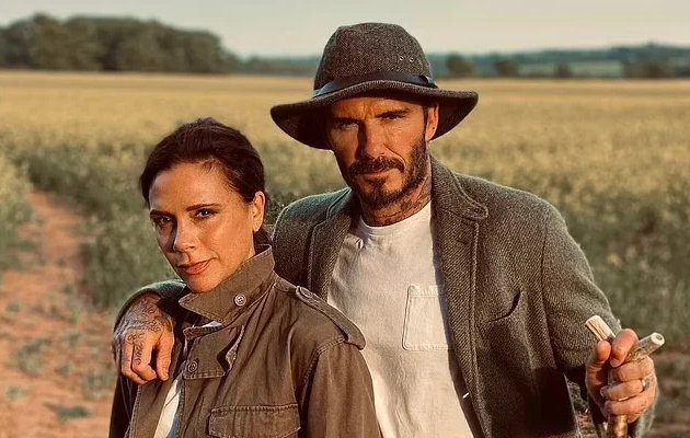 A man and woman in rustic outfits against backdrop of a farm field. Man in hat, leather jacket white t-shirt, woman in brown leather jacket over white t-shirt