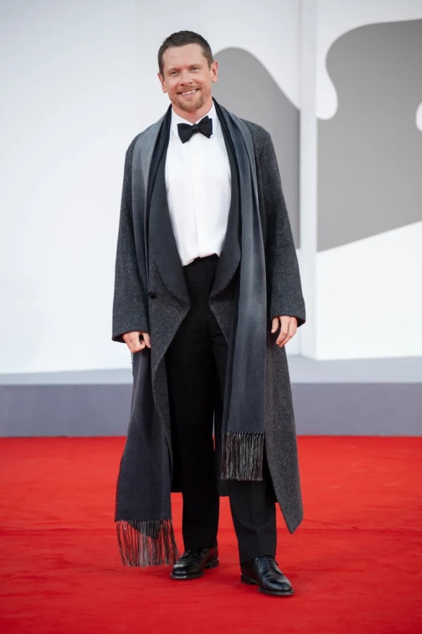 Jack O'Connell cuts a dashing figure on the red carpet at the 83rd Venice Film Festival, wearing a Jonathan Anderson for Dior grey wool overcoat and fringed scarf over a classic black-tie tuxedo.