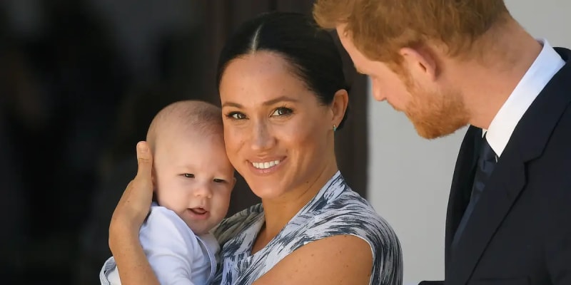 Meghan Markle and Prince Harry smiling together while Harry holds a young girl with long red hair, outdoors in natural light