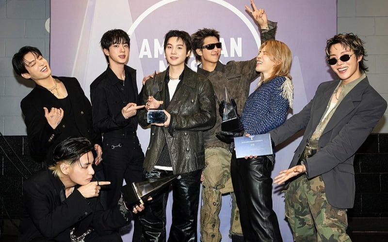 BTS members posing together at a public event, all seven dressed in coordinated outfits and smiling for the camera