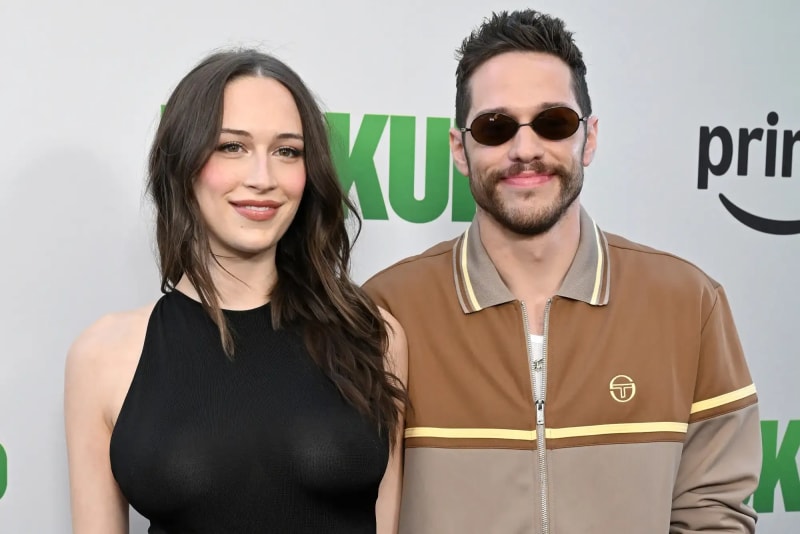 Pete Davidson wearing a grey sweatshirt, white trousers and black sunglasses pushing a baby stroller in a New York City park