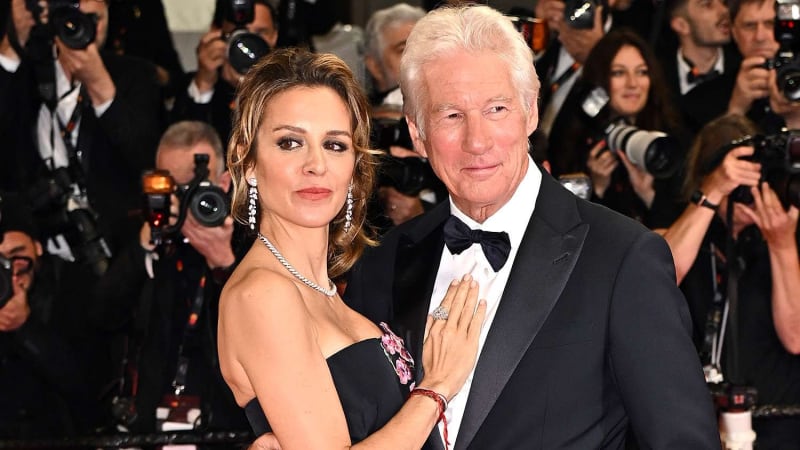 A silver-haired man in casual evening wear stands with a woman in a white maxi dress and block heels at an intimate outdoor birthday gathering, both smiling warmly