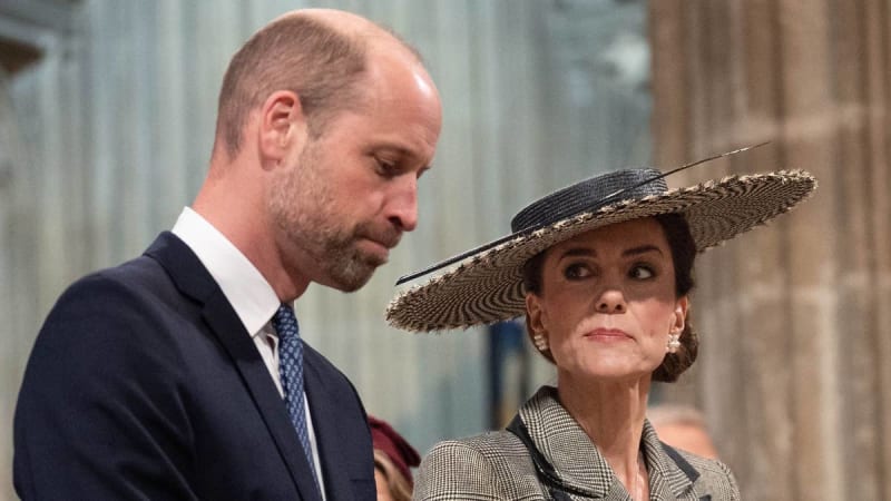 Prince William in black suit bows head with a sombre look as Kate Middleton looks at him with a serious expression, wearing grey coat and large weaved hat