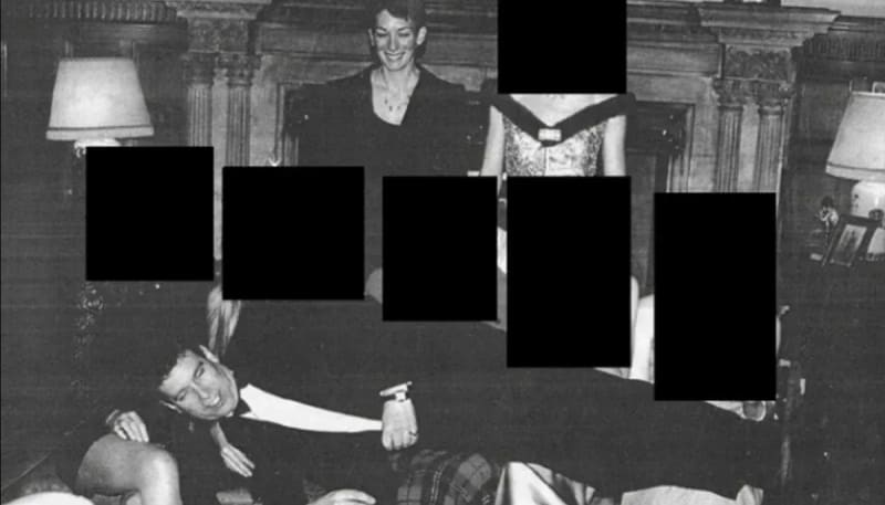 A black-and-white photo of Prince Andrew lying horizontally on his right side on several peoples' laps whose faces are redacted with black rectangles while Ghislaine Maxwell is standing behind looking down smiling, set in an elegant room with a fireplace and lamps.