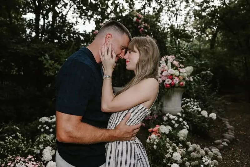 Travis Kelce and Taylor Swift sharing a romantic moment in a lush garden surrounded by pink and white flowers.