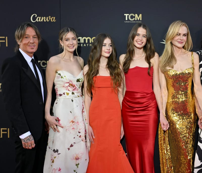 Keith Urban in a black suit, Sunday Rose in a floral gown, Faith Margaret in an orange dress, and Nicole Kidman in a gold sequined gown, posing together on a red carpet backdrop.