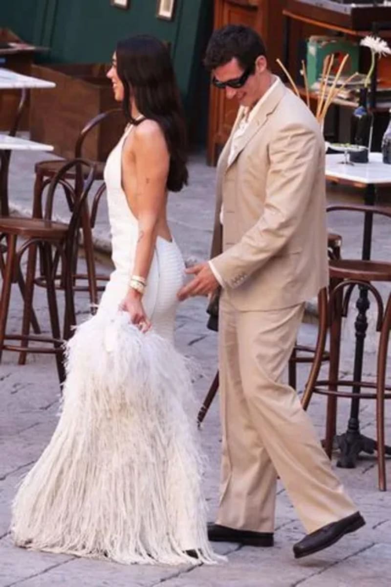 Dua Lipa walking in front of Callum Turner. Lipa is wearing a white dress while Turner wear a cream colored suit