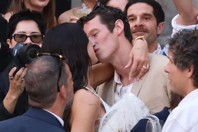 Dua Lipa in whte dress kissed Callum Turner in cream colored suit with people surrounding them