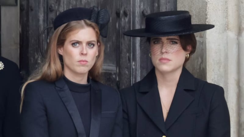 Princesses Beatrice and Eugenie dressed in black mourning attire with black hats, one wearing a veiled fascinator, standing solemnly in front of a large wooden door.