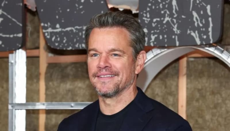 Matt Damon, dark hair and facial hair wearing a black jacket smiles at a formal event.