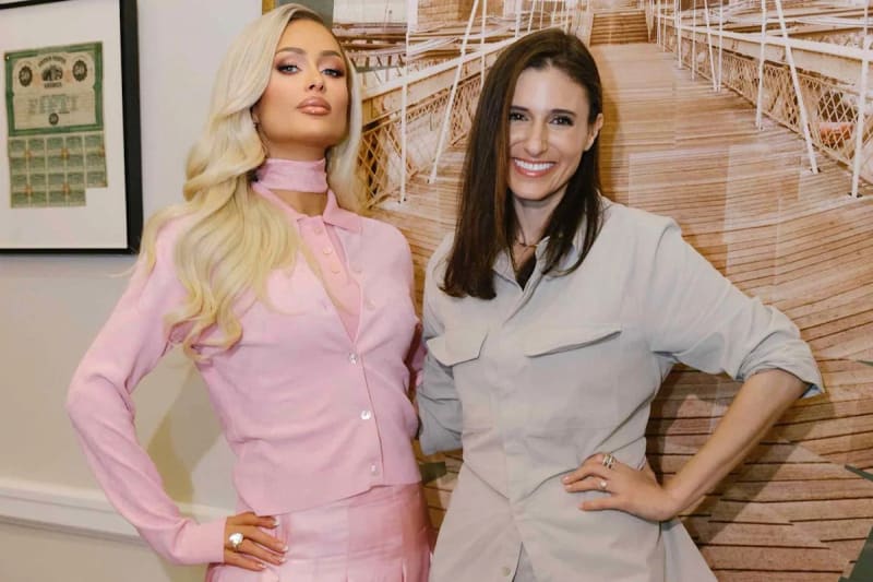 Paris Hilton and Laurie Segall pose together indoors; Paris wears a pink suit with a choker, Segall wears a beige suit, with a wooden boardwalk backdrop.