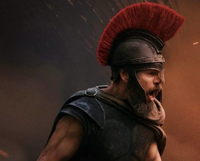 Matt Damon as Odysseus, in ancient Greek armour with a red-plumed helmet, shouting in a dramatic battle scene.