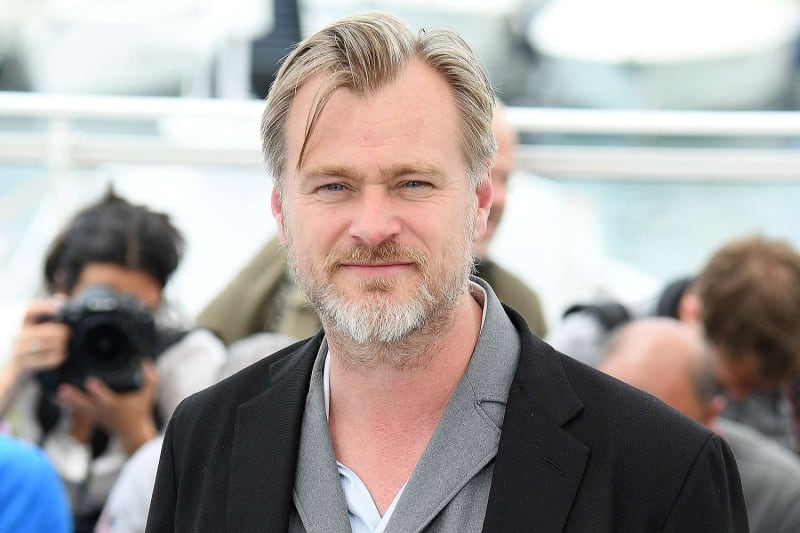 Christopher Nolan in a black blazer and grey shirt at a film event, with photographers visible in the background.
