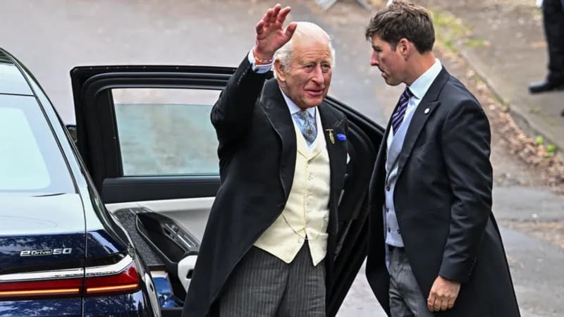 King Charles in wedding guest attire, waving as he steps out of a car at a formal event.