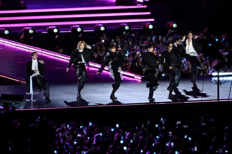 BTS group performs on stage with neon pink lighting while fans in the audience record with their phones.