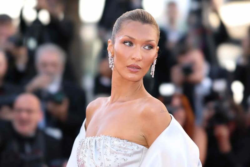 Bella Hadid in elegant white beaded gown and diamond earrings at a formal red carpet event surrounded by photographers.